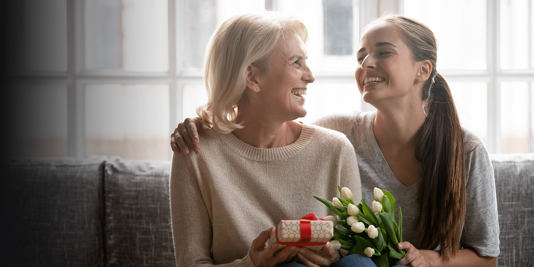 Mother and daughter sitting together smiling on a couch. daughter is giving mother gift of jewelry and tulips