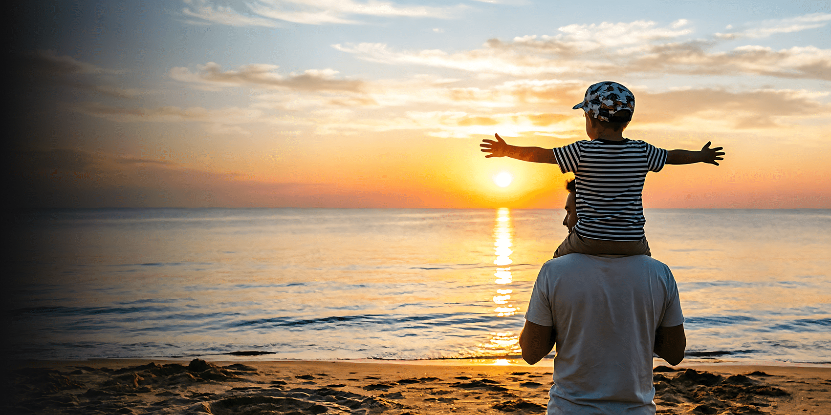 A father standing on a sandy beach facing away, towards the sunset with his young son on his shoulders